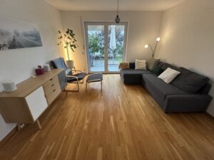 Auf der rechten Seite graue Couch, Fenster bzw. große terrassentüre in der Mitte des Bildes, Schrank links, Parkettboden